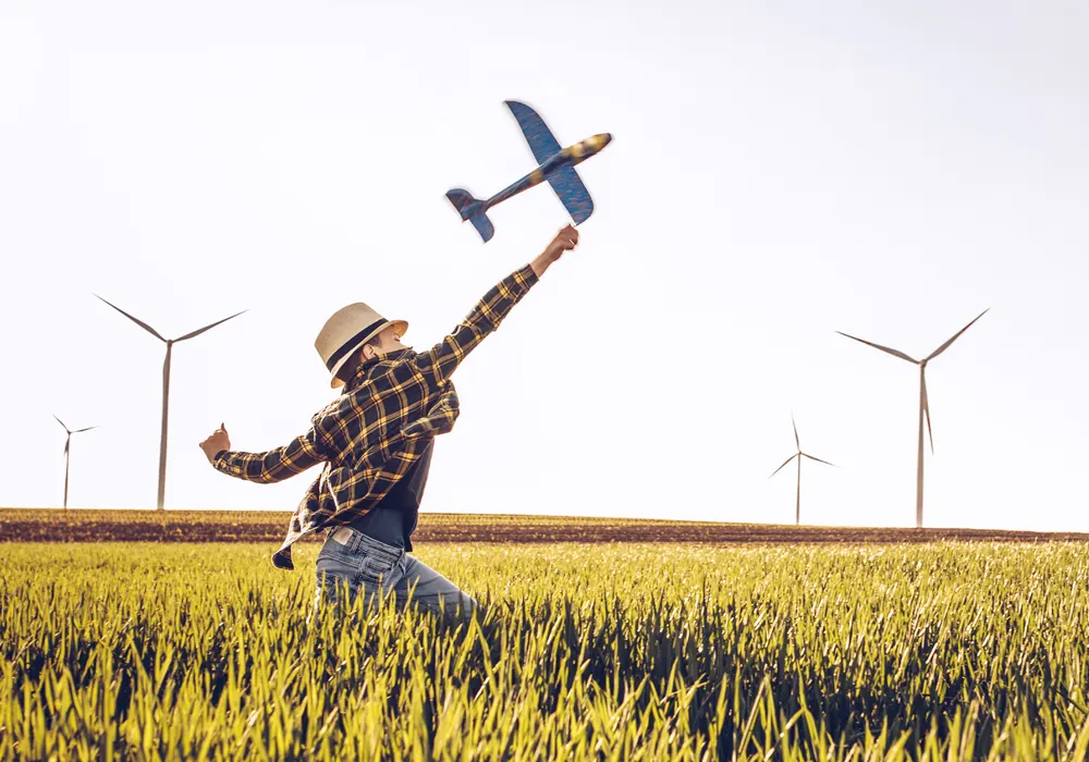 Boy playing with airplane toy in a field with wind turbines in the background.